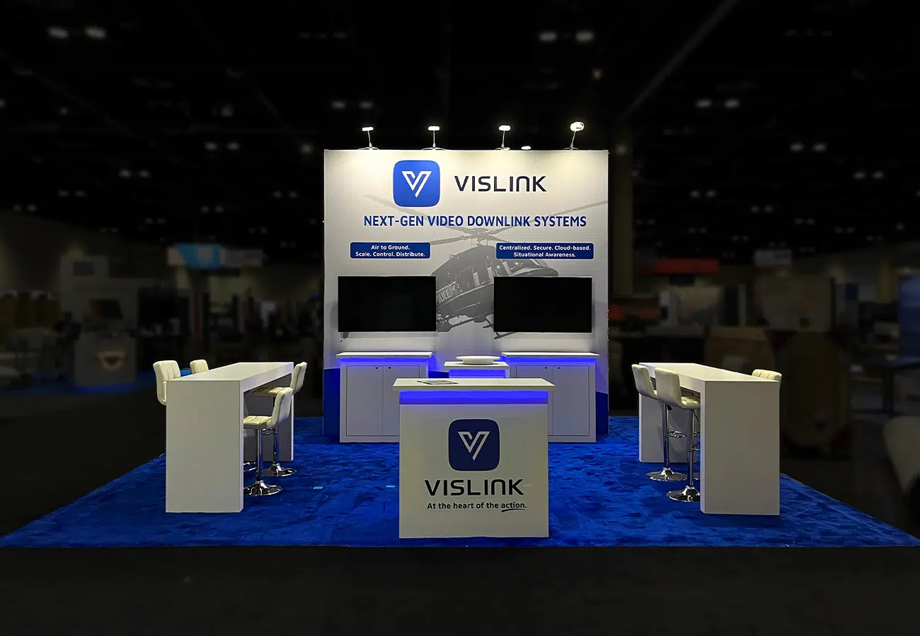 Vislink trade show booth with illuminated branding wall, dual display screens, central reception counter, and blue carpet with seating areas on both sides