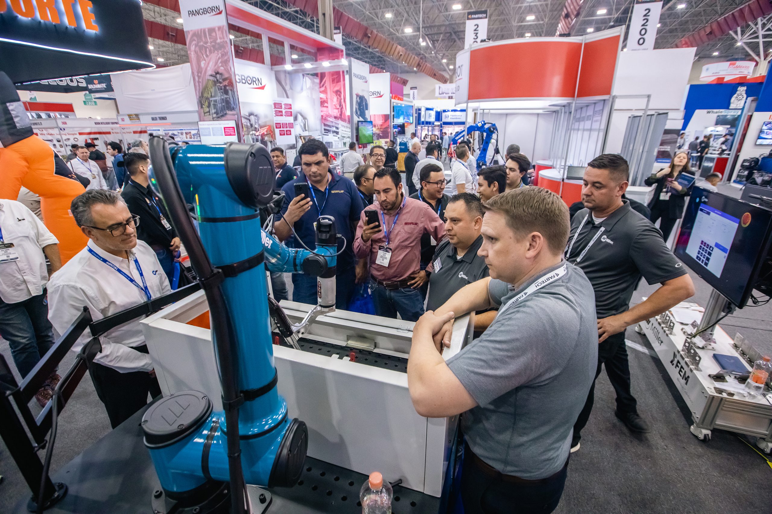 Attendees observing a robotic demonstration at the FABTECH trade show, with various booths and people engaged with the technology.