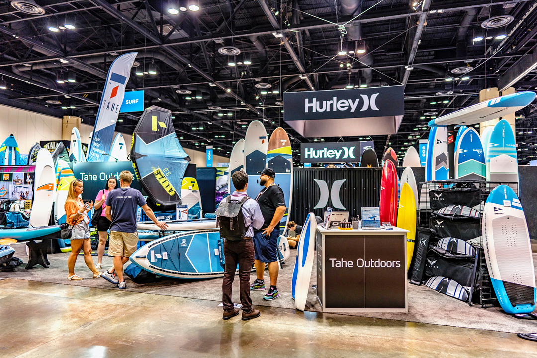 Surf Expo exhibit booth displaying paddle boards, surfboards, and branded signage from Hurley and Tahe Outdoors, with attendees engaging.